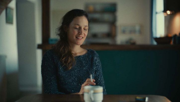 Filmstill aus SERRE MOI FORT. Eine Frau sitzt mit geschlossenen Augen an einem Tisch.