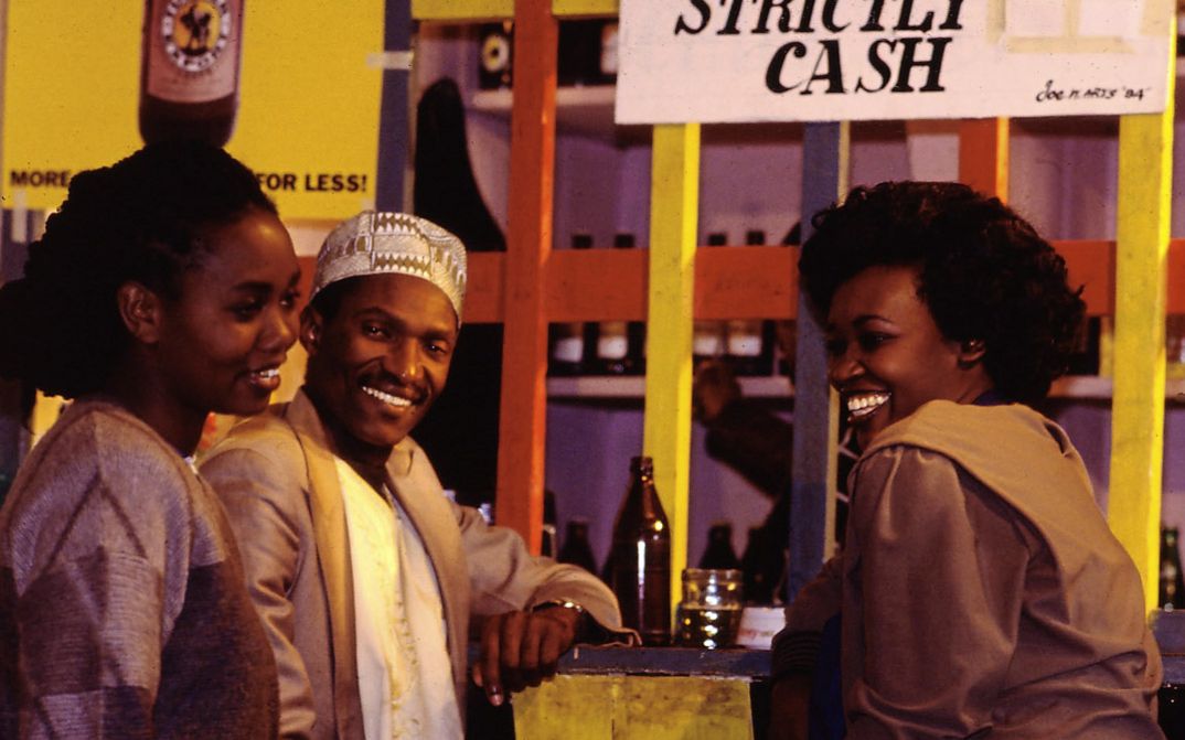 Still from the film „Der Kampf um den heiligen Baum" by Wanjiru Kinyanjui. Two women and a man standing at a bar laughing.