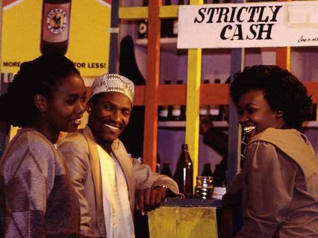 Still from the film „Der Kampf um den heiligen Baum" by Wanjiru Kinyanjui. Two women and a man standing at a bar laughing.