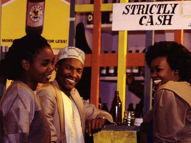 Filmstill aus „Der Kampf um den heiligen Baum“ von Wanjiru Kinyanjui. Zu sehen sind zwei Frauen und ein Mann, die an einer Bar stehen und lachen.