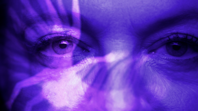 Still from the film "Brainwashed". Close-up of the eyes of a woman who is looking directly into the camera. An abstract image is projected into her face, so that it appears in a blue, purple light.