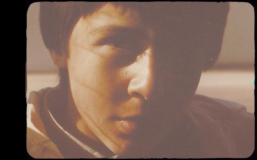 Nahaufnahme eines Jungen, ca. 10-12 Jahre alt, halbes Gesicht im Schatten, warmes Licht, körnige Filmaufnahme.