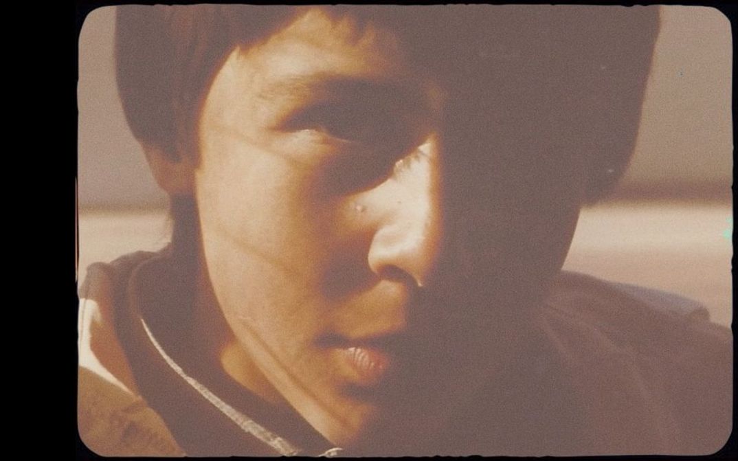 Nahaufnahme eines Jungen, ca. 10-12 Jahre alt, halbes Gesicht im Schatten, warmes Licht, körnige Filmaufnahme.