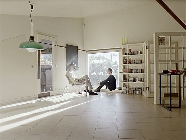 Still from the film "O trio em mi bemol". A man and a woman sit near the window in sparse, spacious room, casting long shadows. 