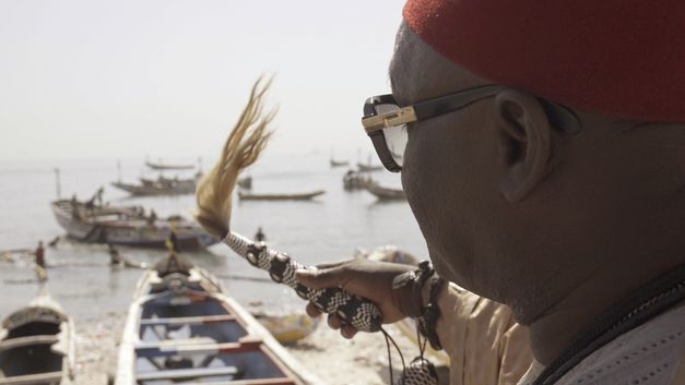 Still from the film "AI: African Intelligence" by Manthia Diawara. A person in the foreground looking at grounded boats on the shore of the lake in the background.