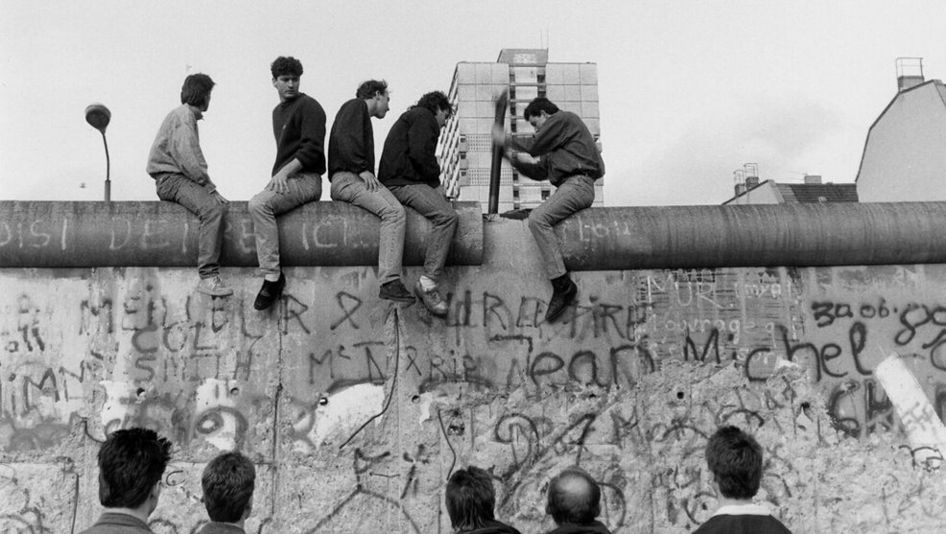 Film still from COUNTDOWN: Some people are sitting on the painted Berlin Wall.