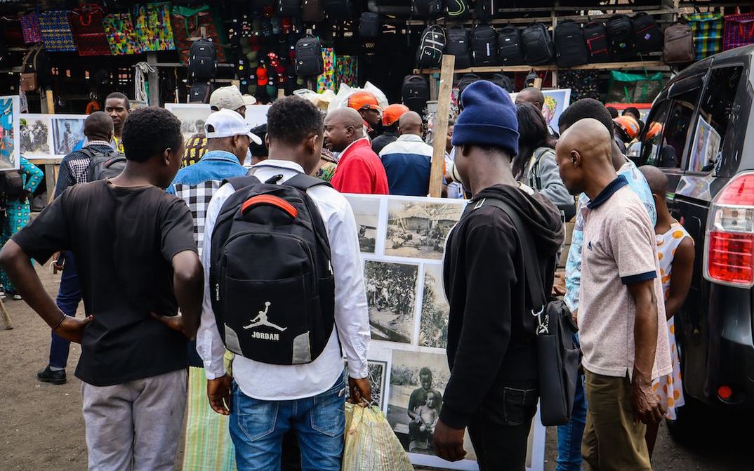 Filmstill aus KUMBUKA: Menschen auf einem Markt, ein paar Männer betrachten eine Fotowand.