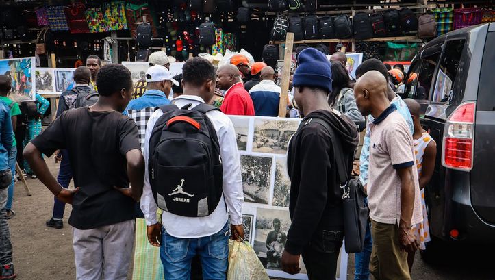 Film still from KUMBUKA: People at a market, a few men looking at a photo wall.