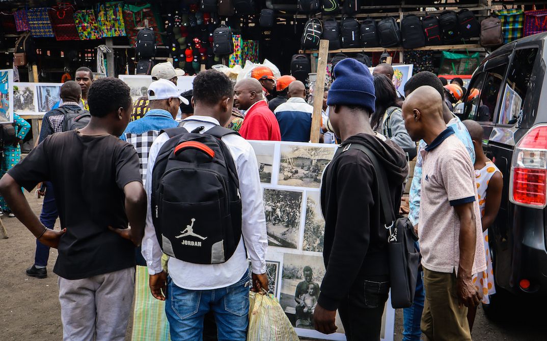Filmstill aus KUMBUKA: Menschen auf einem Markt, ein paar Männer betrachten eine Fotowand.