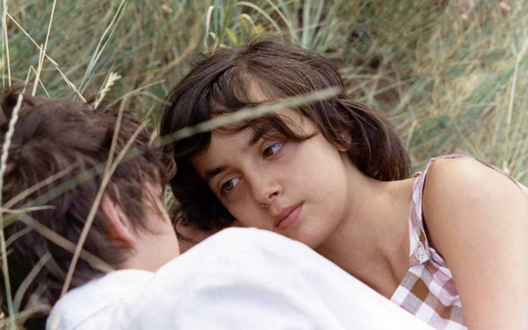 Filmstill aus MES PETITES AMOUREUSES: Ein junges Mädchen liegt in einem Sommerkleid im hohen Gras und blickt auf den Jungen, der ihr gegenüber ist.