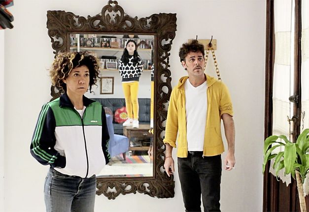 Still from the film "La edad media (The Middle Ages)" by Alejo Moguillansky and Luciana Acuña. A man and a woman stand next to each other in a living room, in the mirror behind them we see the reflection of their daughter standing on a coffee table. 