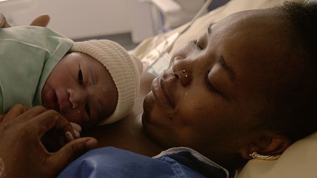 Filmstill aus „Notre corps“ von Claire Simon. Eine Nahaufnahme einer Frau, die liegt und ein Baby an ihrer Brust hält.