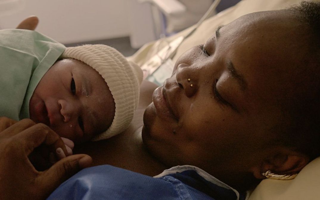 Filmstill aus „Notre corps“ von Claire Simon. Eine Nahaufnahme einer Frau, die liegt und ein Baby an ihrer Brust hält.