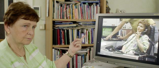 Film still from STOLZ & EIGENSINN by Gerd Kroske. The picture shows a woman pointing at a video on her computer. There is a bookshelf in the background.