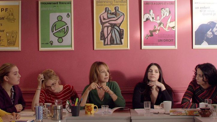 Film still from LES BUREAUX DE DIEU: Five women sit next to each other at a table. Behind them is a pink-painted wall on which several posters about family planning hang.
