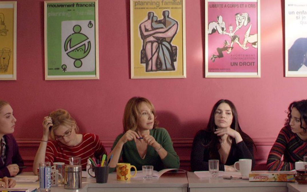 Film still from LES BUREAUX DE DIEU: Five women sit next to each other at a table. Behind them is a pink-painted wall on which several posters about family planning hang.