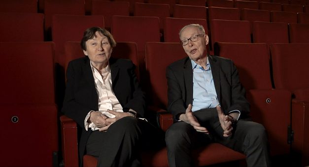 Still from the film "Come With Me to the Cinema – The Gregors" by Alice Agneskirchner. An older woman and a man sitting in a cinema on red seats and looking into the camera. 