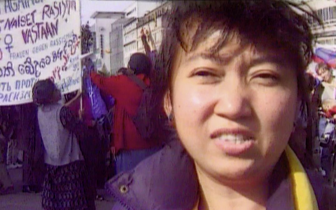 Still from the film "Normality 1–X" by Hito Steyerl. A close-up of a dark-haired woman, a political demonstration in the background with people milling and carrying banners with political slogans. 