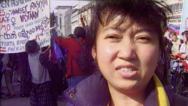 Still from the film "Normality 1–X" by Hito Steyerl. A close-up of a dark-haired woman, a political demonstration in the background with people milling and carrying banners with political slogans. 