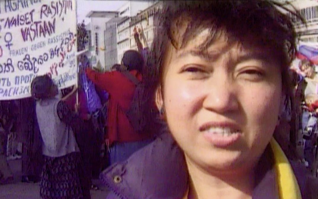 Still from the film "Normality 1–X" by Hito Steyerl. A close-up of a dark-haired woman, a political demonstration in the background with people milling and carrying banners with political slogans.