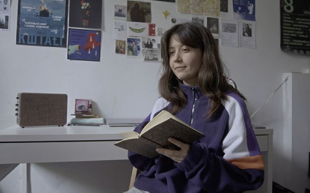 Film still from THE SWAN SONG OF FEDOR OZEROV by Yuri Semashko. It shows a seated young woman with a notebook in her hand and her back to the desk. On the wall are postcards, photos and posters.