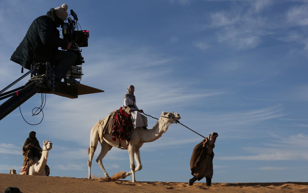 Scene photo from QUEEN OF THE DESERT: A woman on a camel in the desert. In the foreground the film camera with cameraperson.