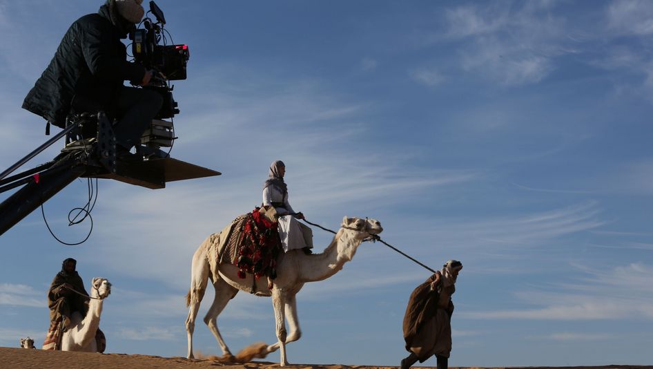 Szenenfoto aus QUEEN OF THE DESERT: Eine Frau auf einem Kamel in der Wüste. Im Vordergrund die Filmkamera mit Kameraperson.