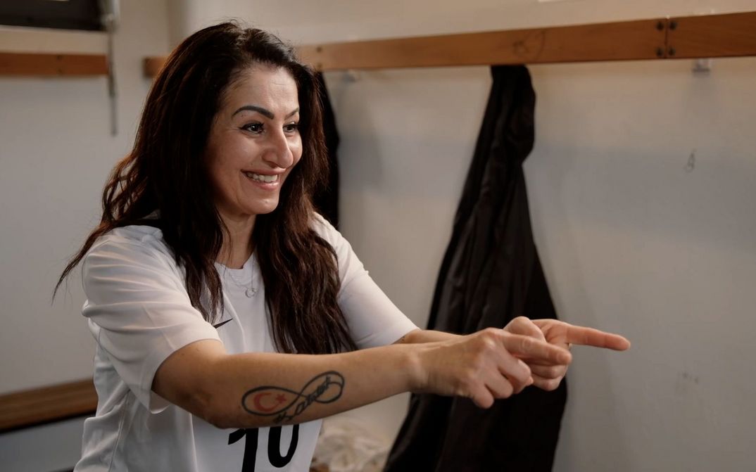Filmstill aus SPIELERINNEN: Eine Frau in einem Fussballtrikot steht in einer Umkleidekabine und motiviert ihre Mitspielerinnen, die nicht im Bild sind.