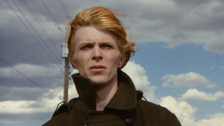 Filmstill aus THE MAN WHO FELL TO EARTH: David Bowie blickt mit windzerzausten Haaren in die Ferne. Man sieht den wolkigen Himmel und Strommasten und Stromkabel.
