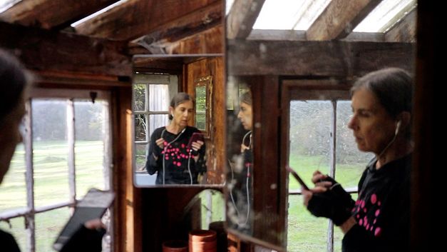 Filmstill aus dem Film "Horse Opera" von Moyra Davey. Eine Frau links im Bild spricht über weiße Kopfhörer in ihr Handy. Auf der rechten Seite und im Hintergrund sind zwei Spiegel, sodass die Frau dreimal im Bild zu sehen ist.