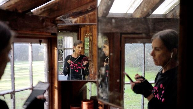 Still from the film "Horse Opera" by Moyra Davey. A women on the left side of the frame is speaking into her phone through white earphones. There are mirrors on the right side and in the background, so that the woman can be seen thrice in the picture.