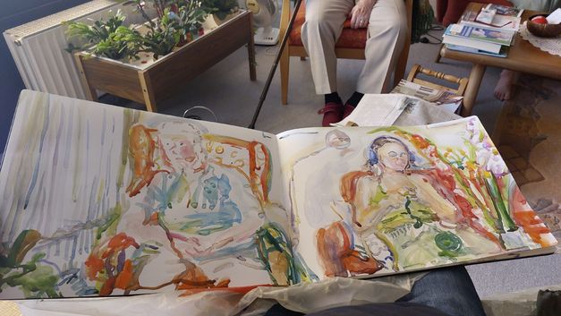 Film still from 2024 (2023) by Stefan Hayn. The picture shows an open sketchbook on a lap. A person with a crutch is sitting in the background.