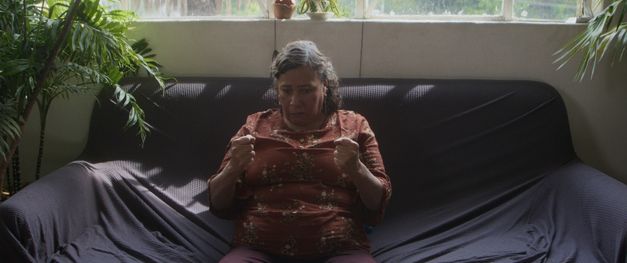 Eine Frau mittleren Alters sitzt allein auf Sofa, Hände angespannt, Licht von Fenster.