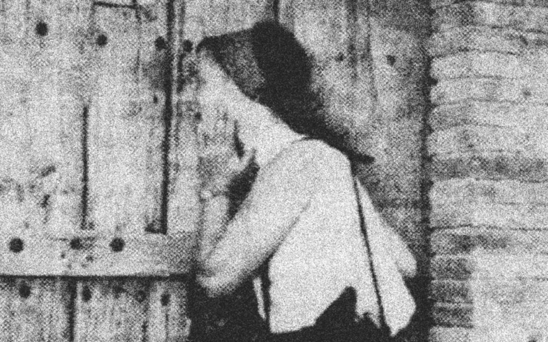 Filmstill aus dem Film „Exhibition“ von Mary Helena Clark. Eine Person an einer Holztür; das Bild ist schwarz-weiß und stark gekörnt.