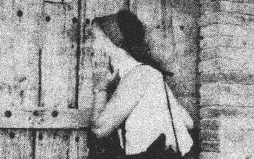 Film still from Mary Helena Clark’s „Exhibition“. A person at a wooden door; the picture is in black and white and very grainy.