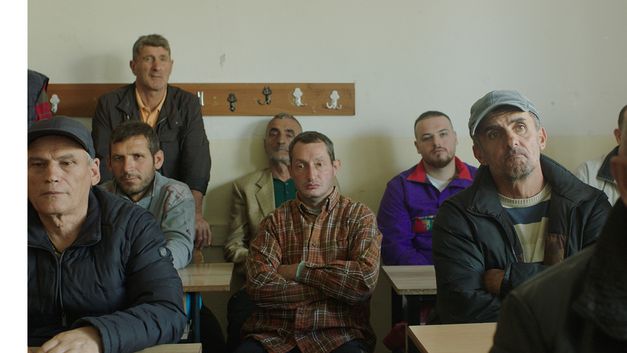 Film still from Alban Muja’s film “Mua besoj më shpëtoj portreti”. A classroom is filled with men of various ages facing the camera.