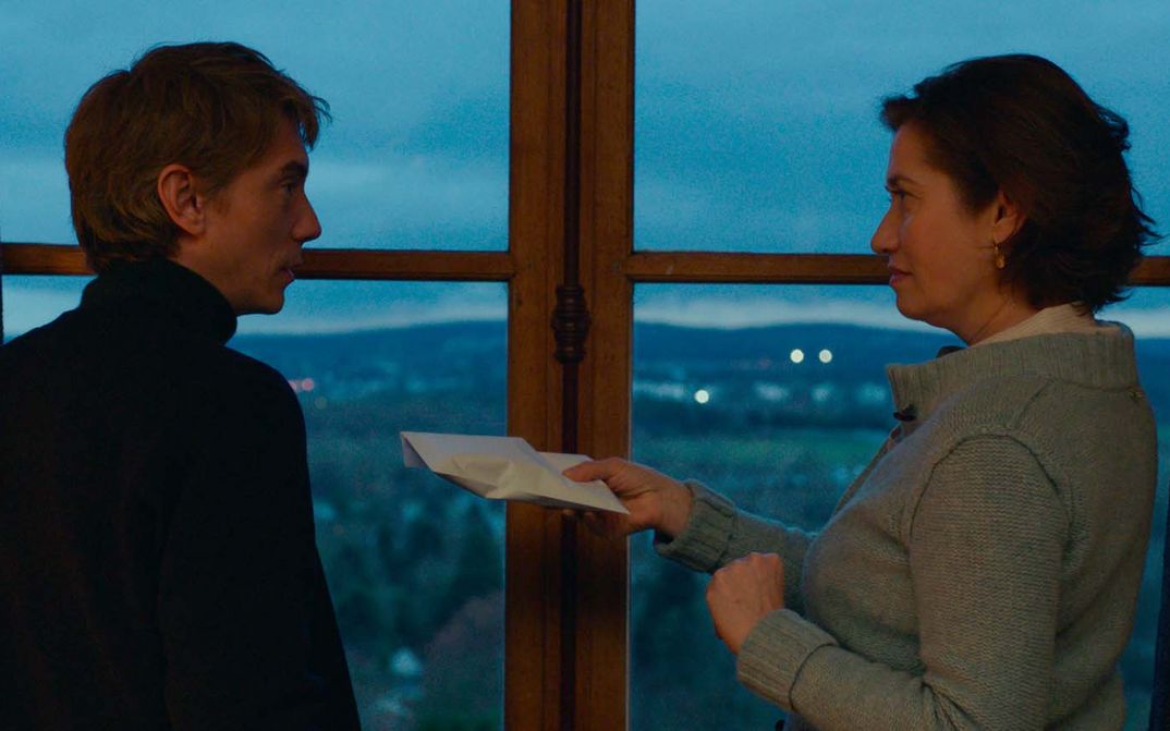 Filmstill from VOUS NE DESIREZ QUE MOI: Two people are standing in front of the window. The person on the right is holding an envelope out to the other person.