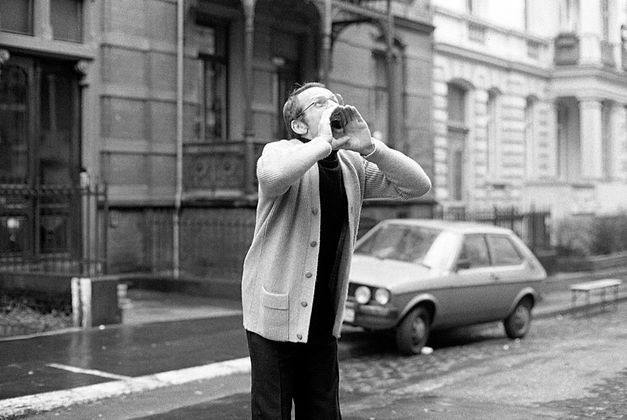 Black and white image of a man in a cardigan standing on the street. He has put his hands to his mouth to shout. In the background there is a car on the pavement. 