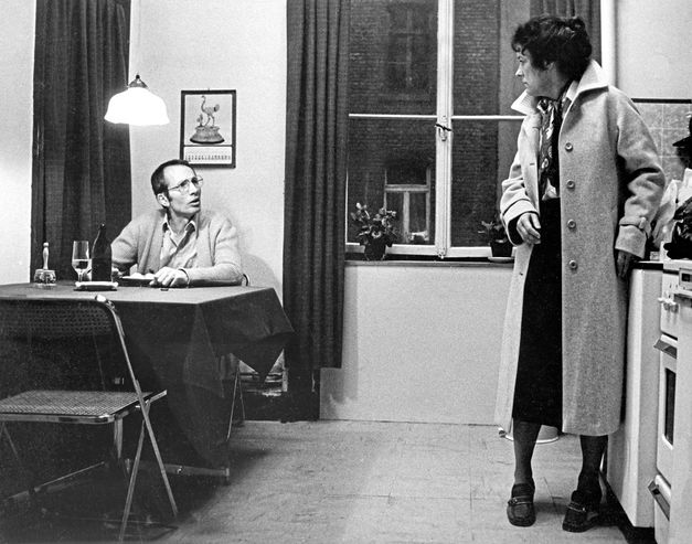 Black and white image of a man wearing glasses and a woman in a coat in a kitchen. The man is sitting at a table with a plate of food and a beer with a glass. The woman is standing at the kitchen counter. They are looking at each other and talking.