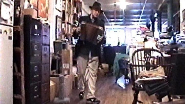 Film still from NOTES ON UTOPIA. Jonas Mekas with an accordion in a crowded office.