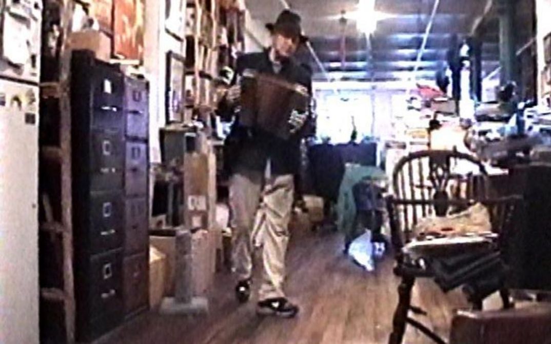 Filmstill aus NOTES ON UTOPIA. Jonas Mekas mit einer Ziehharmonika in einem vollgestellten Büro.