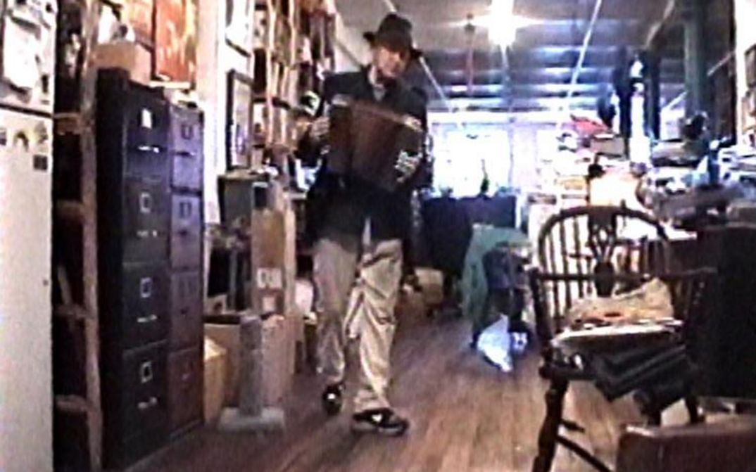 Filmstill aus NOTES ON UTOPIA. Jonas Mekas mit einer Ziehharmonika in einem vollgestellten Büro.