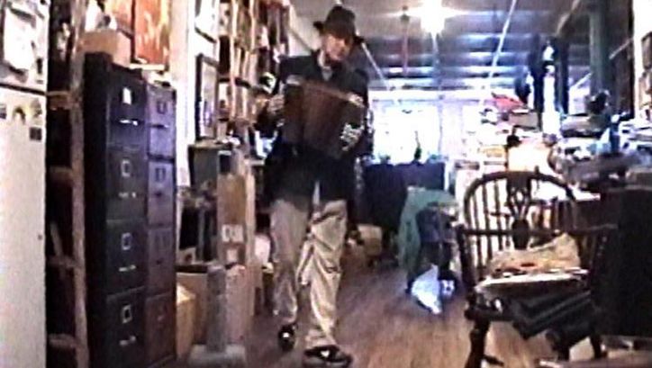 Filmstill aus NOTES ON UTOPIA. Jonas Mekas mit einer Ziehharmonika in einem vollgestellten Büro.