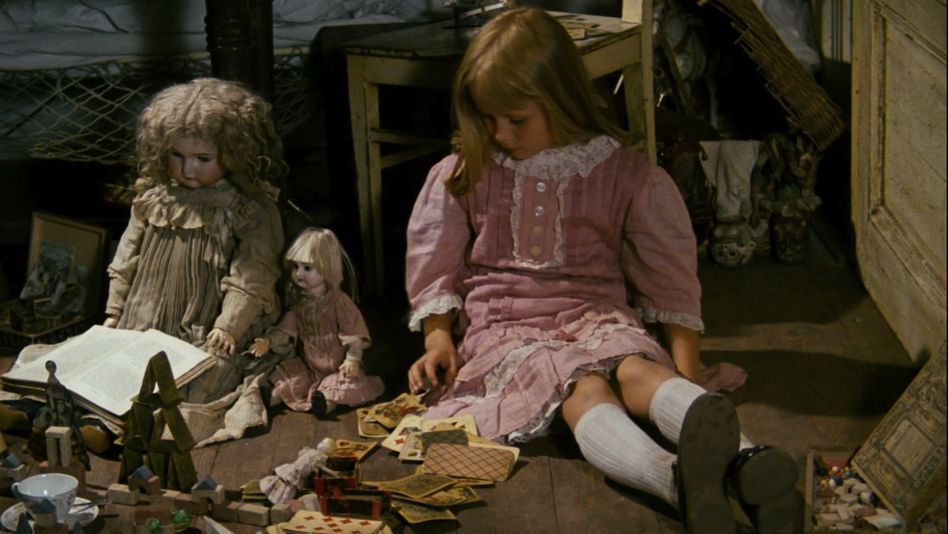Film still from ALICE: A blonde girl in a pink dress sits on the floor next to dolls and other toys.