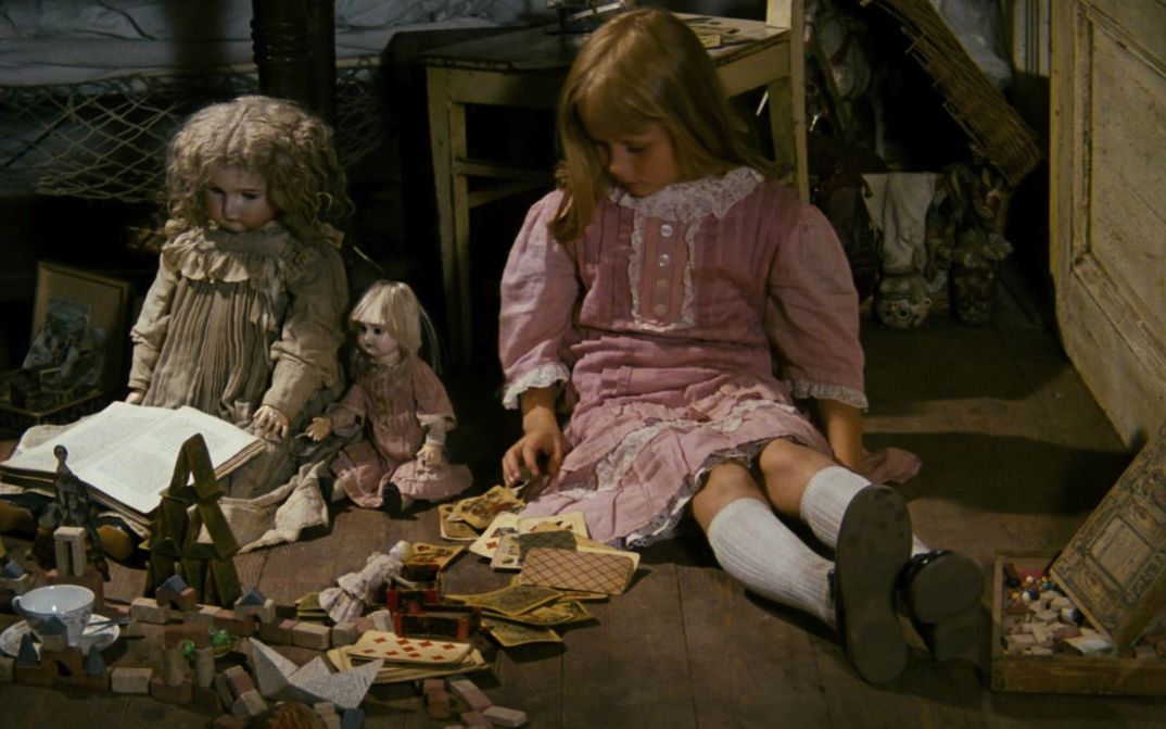 Filmstill aus ALICE: Ein blondes Mädchen in einem rosafarbenen Kleid sitzt neben Puppen und weiterem Spielzeug auf dem Boden.