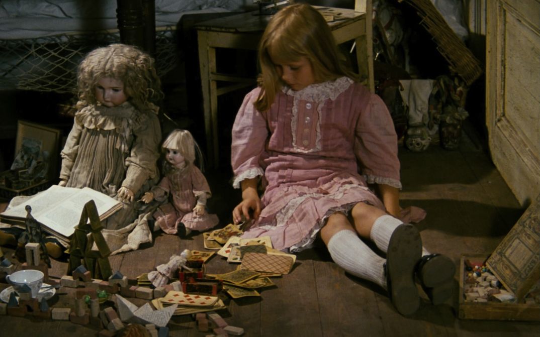 Filmstill aus ALICE: Ein blondes Mädchen in einem rosafarbenen Kleid sitzt neben Puppen und weiterem Spielzeug auf dem Boden.