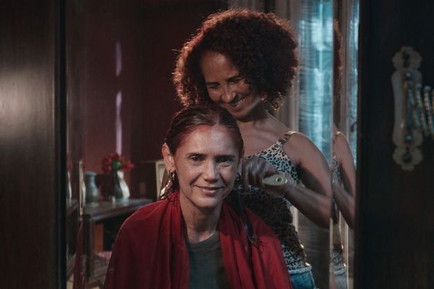 Zwei Frauen stehen in einem Innenraum, eine sitzt auf einem Stuhl, während die andere ihr die Haare kämmt.