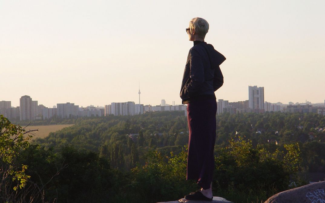Filmstill aus THE INVISIBLE FRAME: Tilda Swinton schaut aus einer leicht erhöhten Position auf eine Reihe von Hochhäusern, im Hintergrund ist der Berliner Fernsehturm zu erkennen.