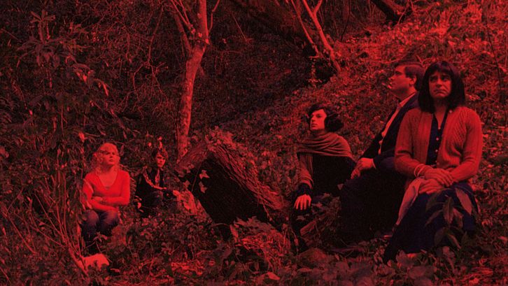 Film still from MORRER COMO UM HOMEM: A red coloured image of a group of people sitting in a forest.