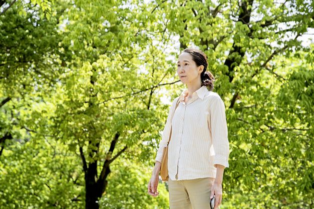 Zu sehen ist eine Frau mit dunklen, zu einem Zopf gebundenen Haaren und einem weißen Hemd. Sie schaut durch die Gegend. Im Hintergrund stehen hellgrüne Bäume.